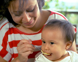 A mother feeds her baby complementary food with a spoon as he sits on her lap.